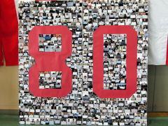 一人ずつ胸の前に紙を広げた写真を何枚も並べた中央に大きな数字で80の赤文字で80周年を記念して作られた作品の写真